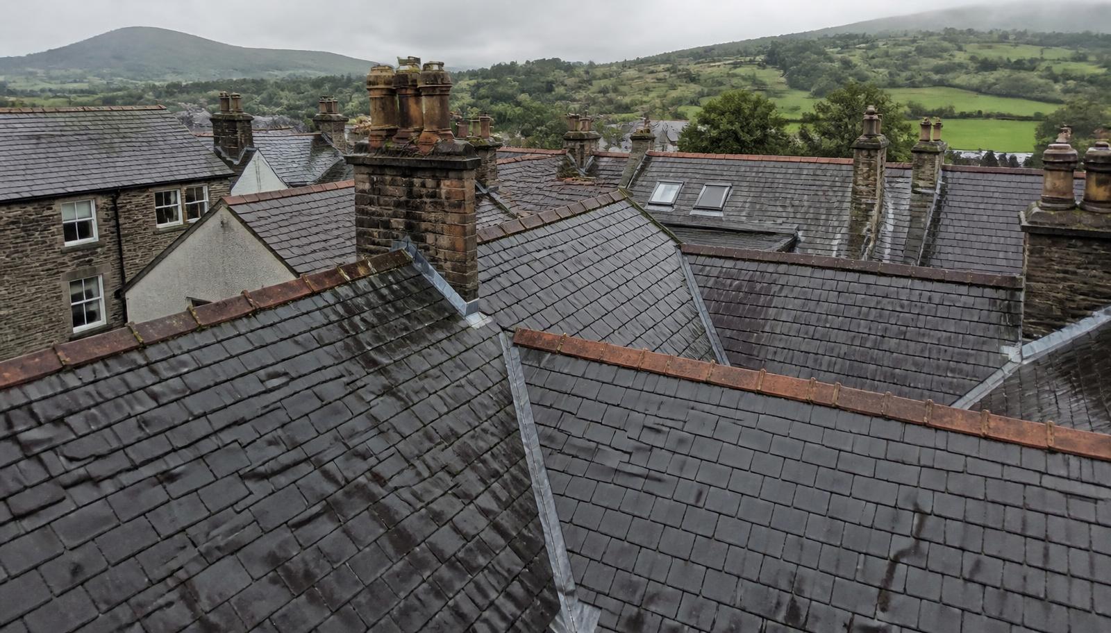 Survey drone hovering above a slate roof in Cumbria on an overcast day, photographing the ridge and chimney stack